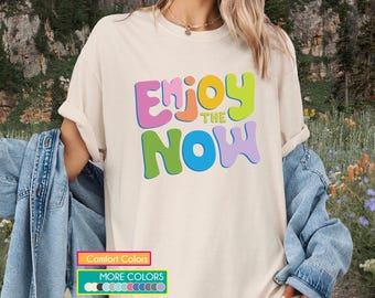 May include: Cream-colored t-shirt with the words "Enjoy the Now" in colorful, bubble-style letters. The shirt is part of a clothing collection, with a variety of color options available. The shirt is worn with a denim jacket.