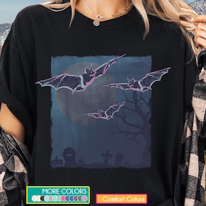 May include: Black t-shirt featuring a Halloween-themed graphic. The design includes bats in flight against a moonlit sky, a graveyard scene, and a tree silhouette. The shirt has a crew neck and short sleeves.