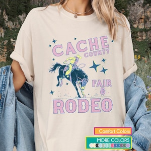 May include: Cream-colored t-shirt featuring a rodeo design. The graphic includes the text "CACHE COUNTY FAIR & RODEO" in pink and blue, along with a cowboy on a bucking bronco. Additional text at the bottom indicates "Comfort Colors" and "More Colors".