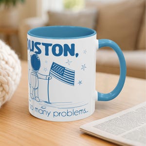 May include: White ceramic mug with a blue interior and handle. The mug features a blue illustration of an astronaut holding an American flag, with the text "HOUSTON, so many problems..." in blue.
