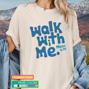 Könnte beinhalten: Cremefarbenes T-Shirt mit dem Text "Walk With Me" in blauen Buchstaben, umrandet von Rosa und Gelb. Der Text "Moses 6:34" steht darunter. Das Shirt wird mit einer Jeansjacke getragen.