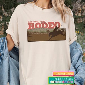 Pode incluir: Camiseta creme com um gráfico de rodeio. O gráfico inclui o texto "Cache County Fair and Rodeo" acima de uma silhueta de um cavaleiro em um cavalo selvagem. A camisa também tem texto "Comfort Colors" e "More Colors".
