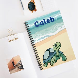 May include: A blue and green spiral notebook with a cartoon sea turtle on a beach with waves and the name "Caleb" printed on the cover.