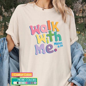 Könnte beinhalten: Cremefarbenes T-Shirt mit dem Text "Walk With Me" in bunten Buchstaben. Der Text enthält "Moses 6:34". Das T-Shirt ist Teil einer Bekleidungskollektion.