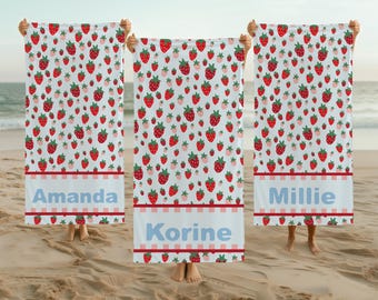 May include: Beach towels with a white background and a pattern of red strawberries. Each towel has a personalized name in a light blue font. The towels are displayed on a sandy beach.