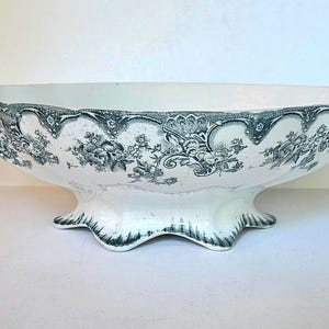 May include: An oval ceramic serving bowl featuring a white base with a dark green floral transferware pattern. The bowl has a scalloped rim, two decorative handles on the sides, and a pedestal base with a matching dark green floral design.