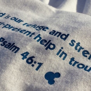 May include: White t-shirt with blue text that reads "God is our refuge and strength, an ever-present help in trouble. Psalm 46:1"