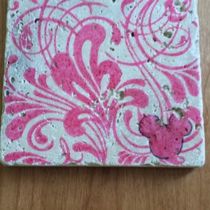 May include: A square, white stone coaster with a pink floral design and a pink Mickey Mouse silhouette.