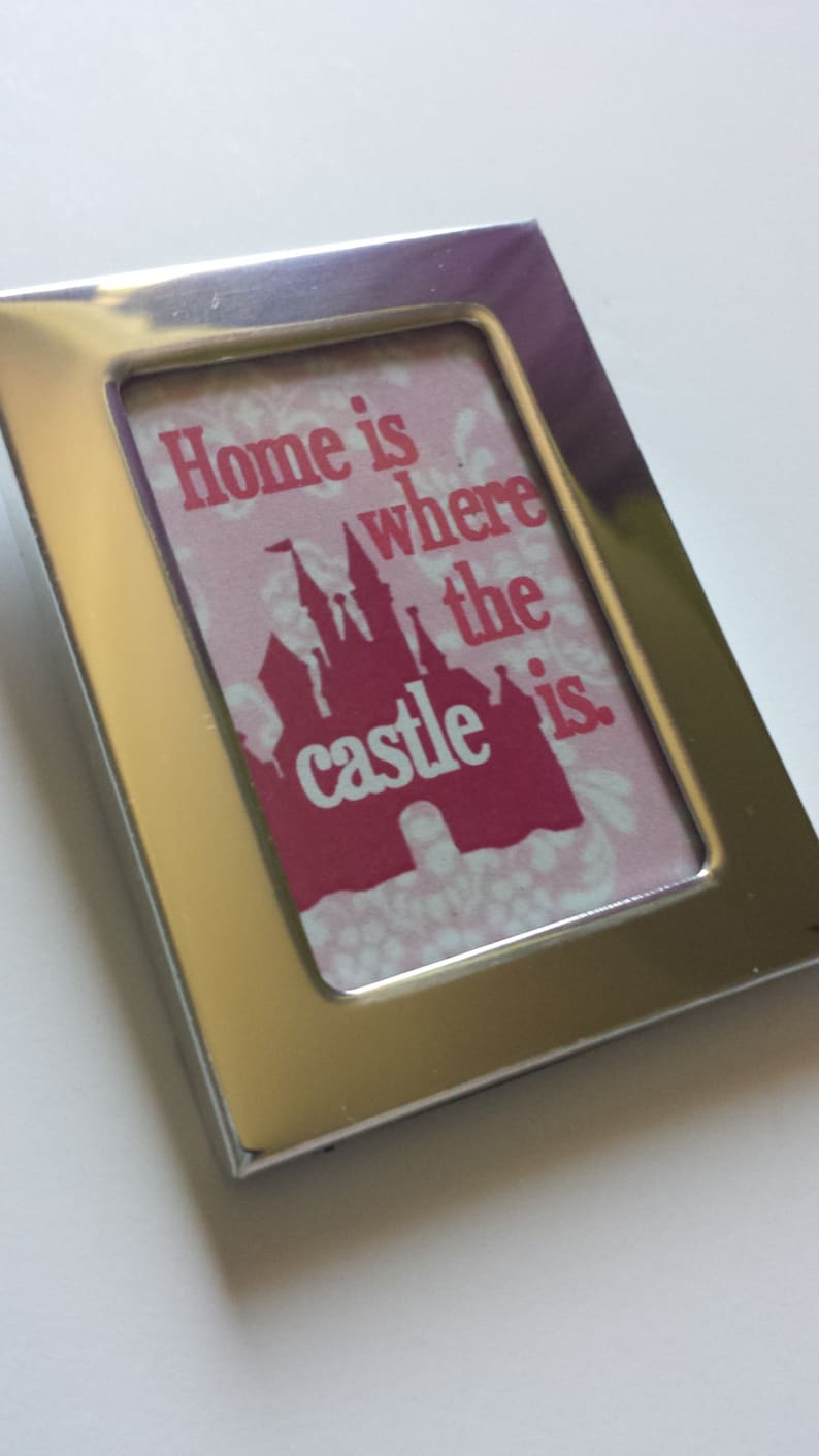 May include: A silver picture frame with a pink and red print inside. The print features a silhouette of a castle and the text "Home is where the castle is."