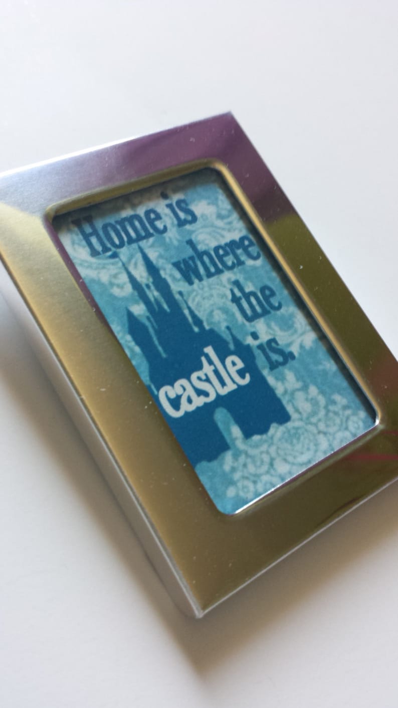 May include: A silver picture frame with a blue and white image of a castle and the text "Home is where the castle is."