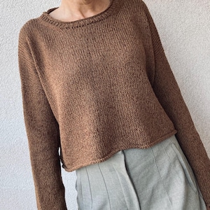 Könnte beinhalten: Ein brauner Strickpullover mit Rundhalsausschnitt und langen Ärmeln. Der Pullover hat einen leicht verkürzten Saum. Das Model trägt eine graue Hose. Der Pullover ist aus einem strukturierten Strickstoff gefertigt.