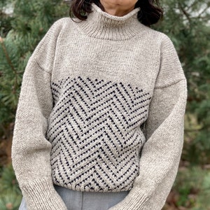 Puede incluir: Un su&eacute;ter beige de cuello alto con un patr&oacute;n de chevron blanco y negro en la parte delantera. El su&eacute;ter est&aacute; hecho de una tela suave y tejida.