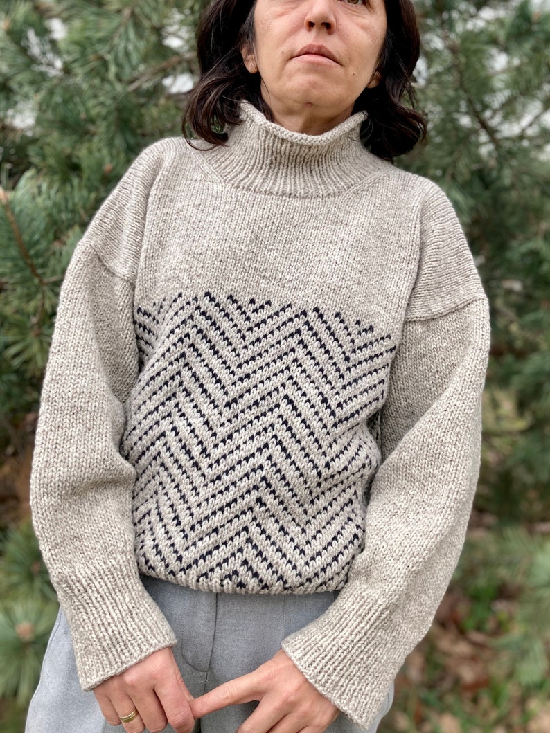 Puede incluir: Un su&eacute;ter beige de cuello alto con un patr&oacute;n de chevron blanco y negro en la parte delantera. El su&eacute;ter est&aacute; hecho de una tela suave y tejida.