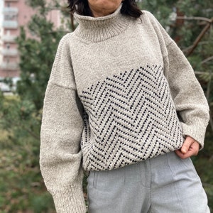 Puede incluir: Un su&eacute;ter beige de cuello alto con un patr&oacute;n de chevron blanco y negro en la parte delantera. El su&eacute;ter tiene mangas largas y un ajuste holgado.