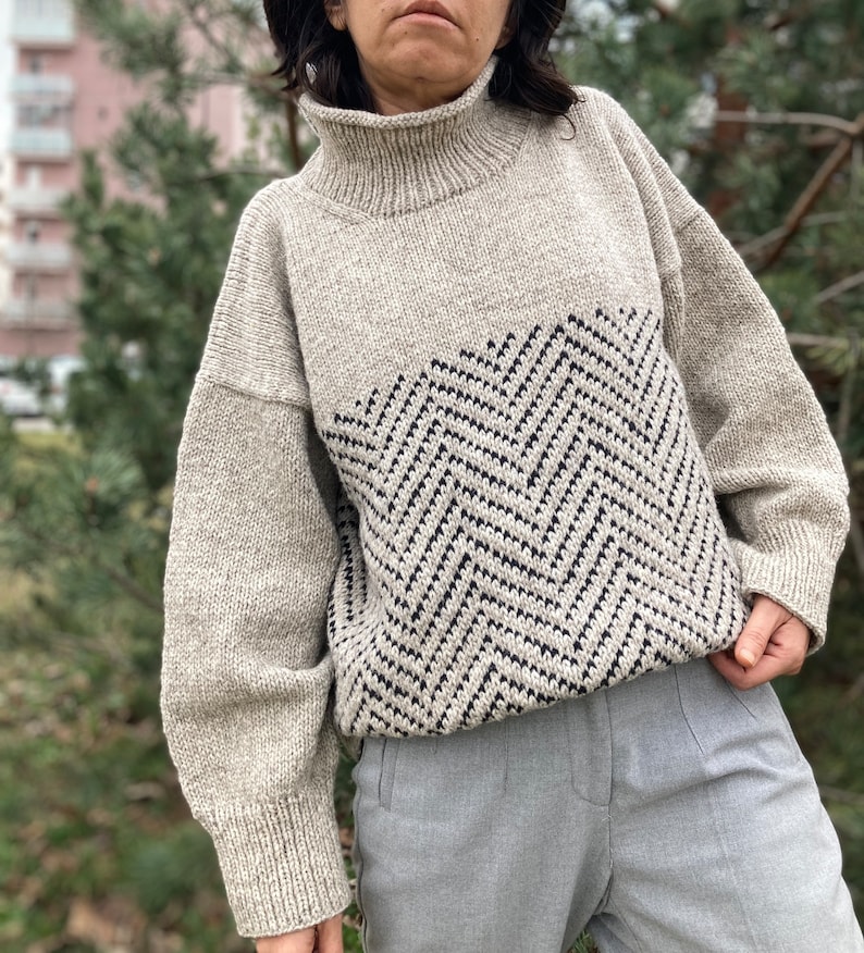 Puede incluir: Un su&eacute;ter beige de cuello alto con un patr&oacute;n de chevron blanco y negro en la parte delantera. El su&eacute;ter tiene mangas largas y un ajuste holgado.
