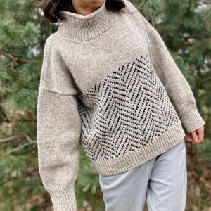 Puede incluir: Un su&eacute;ter de punto beige con un patr&oacute;n de chevron blanco y negro en la parte delantera. El su&eacute;ter tiene cuello alto y mangas largas.