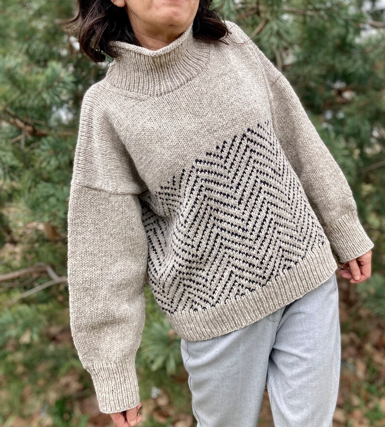 Puede incluir: Un su&eacute;ter de punto beige con un patr&oacute;n de chevron blanco y negro en la parte delantera. El su&eacute;ter tiene cuello alto y mangas largas.