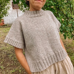 May include: A light brown knitted sweater with a high neckline and short sleeves. The sweater has a textured knit and a slightly cropped length.