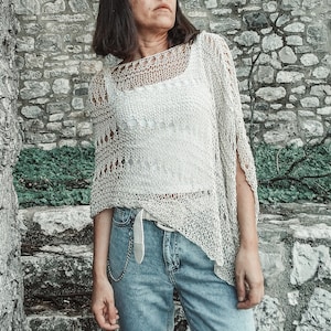 Puede incluir: Un top blanco de crochet con un diseño suelto y tejido abierto. El top se lleva sobre un top blanco de tirantes y se combina con unos vaqueros azules.