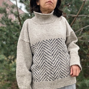 Puede incluir: Un su&eacute;ter beige de cuello alto con un patr&oacute;n de chevron en negro y gris oscuro. El su&eacute;ter tiene un corte holgado y mangas largas.
