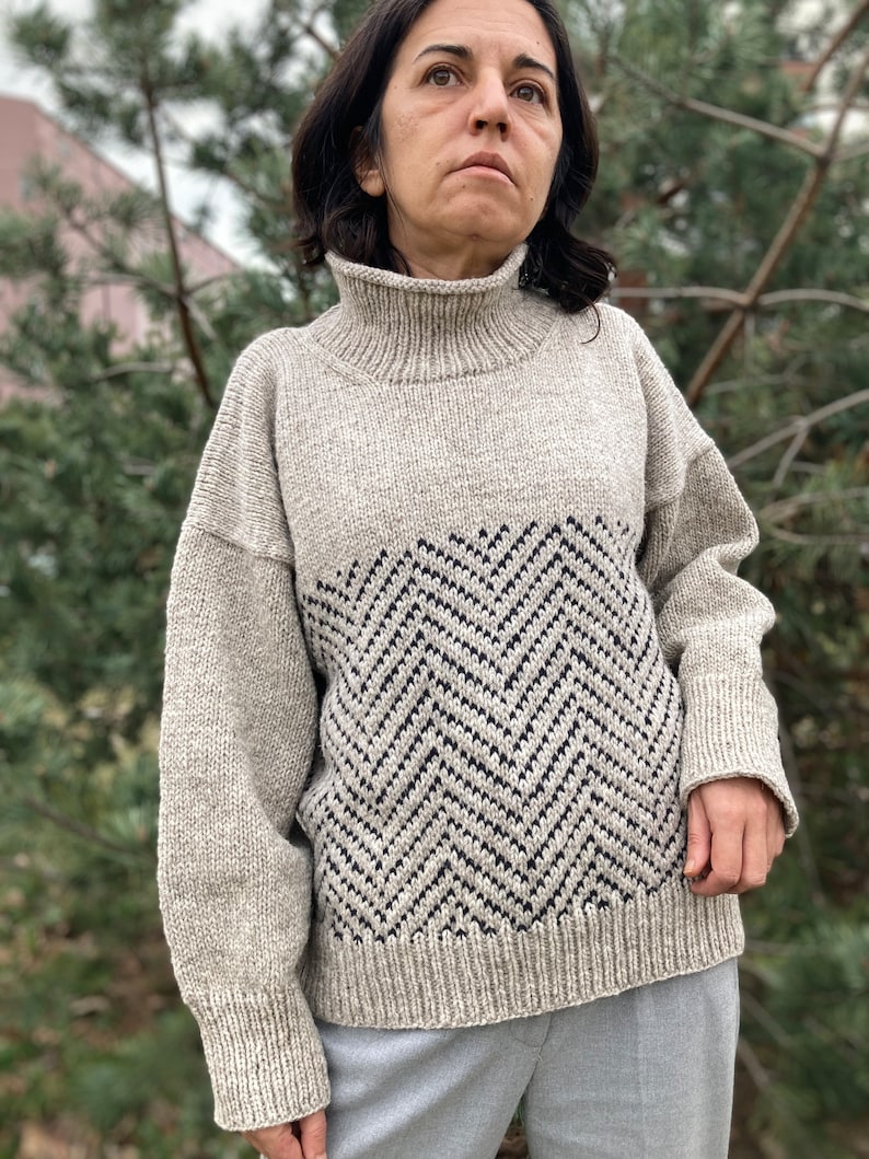 Puede incluir: Un su&eacute;ter beige de cuello alto con un patr&oacute;n de chevron en negro y gris oscuro. El su&eacute;ter tiene un corte holgado y mangas largas.