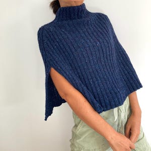 May include: A navy blue knitted poncho with a high neck and ribbed texture. The poncho has slits on the sides and a cropped length.