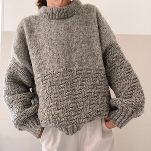 May include: A gray knitted sweater with a textured pattern on the bottom half. The sweater has a high neck and long sleeves.