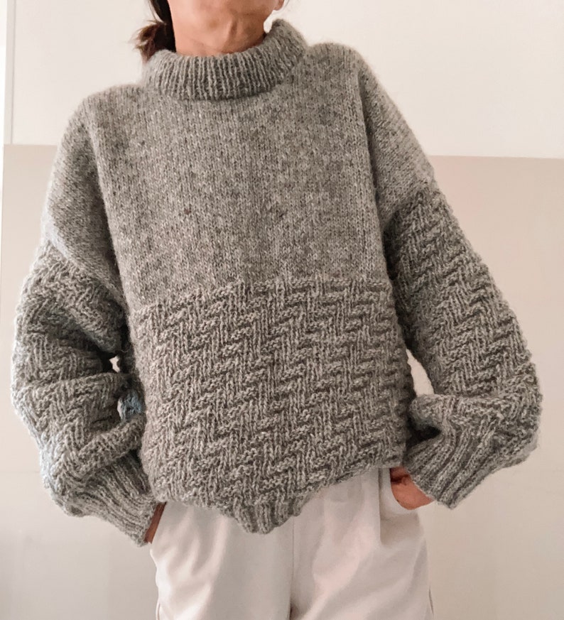 May include: A gray knitted sweater with a textured pattern on the bottom half. The sweater has a high neck and long sleeves.