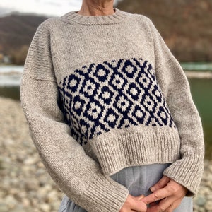 Op de afbeelding: Een beige gebreide trui met een geometrisch patroon in marineblauw. De trui heeft een getailleerde lengte en lange mouwen.