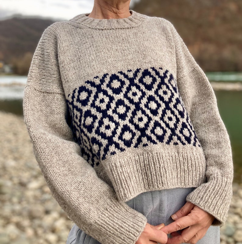 Op de afbeelding: Een beige gebreide trui met een geometrisch patroon in marineblauw. De trui heeft een getailleerde lengte en lange mouwen.