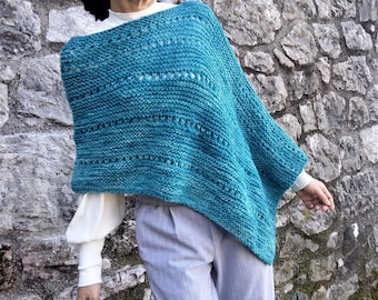 May include: A teal blue knitted poncho with a lacy pattern. The poncho is worn by a person standing in front of a stone wall.