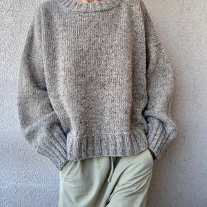 May include: A gray knitted sweater with a round neckline and long sleeves. The sweater has a speckled texture and a relaxed fit.