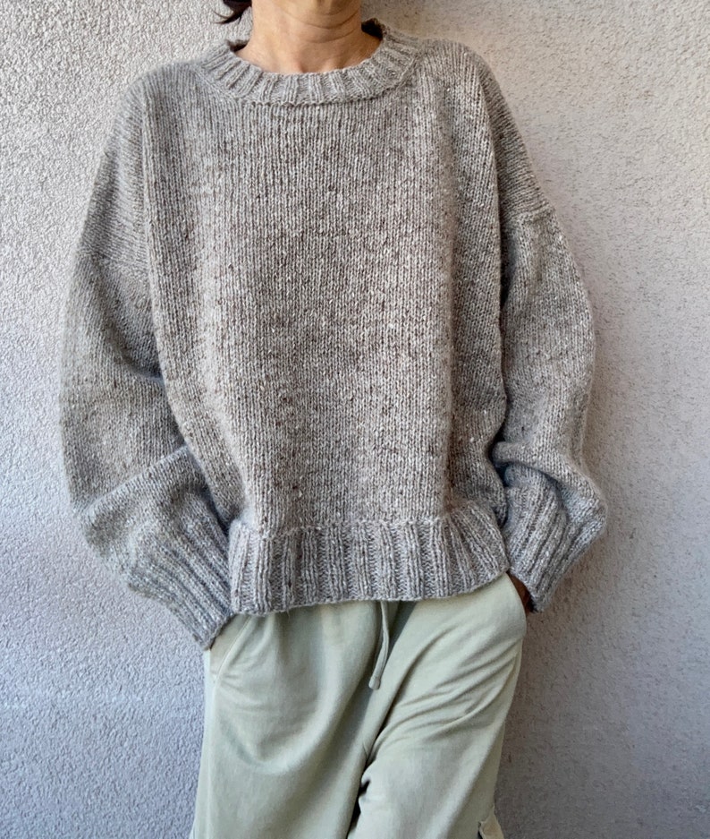 May include: A gray knitted sweater with a round neckline and long sleeves. The sweater has a speckled texture and a relaxed fit.