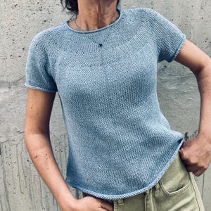 May include: A pale blue knitted t-shirt with short sleeves and a round neckline. The fabric has a textured appearance. The hemline is slightly curved. The t-shirt is a solid colour and has a simple, classic design, suitable for casual wear.