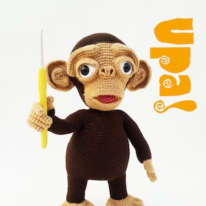 May include: A crocheted monkey toy holding a yellow crochet hook. The monkey is brown with tan fur and has large, round eyes. The toy is standing on a white background.