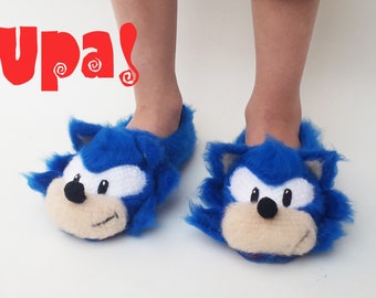 May include: Blue fuzzy slippers with a Sonic the Hedgehog face on the front. The slippers are worn by a person's feet. The text "Upa!" is written in red at the top of the image.