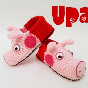 Puede incluir: Un par de pantuflas de crochet rosa y rojo con forma de cara de cerdo. Las pantuflas tienen ojos negros y una boca cosida negra. El texto "Upa!" está escrito en letras rojas sobre un fondo blanco.
