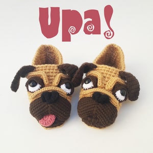 Puede incluir: Un par de pantuflas de crochet hechas a mano con forma de perro pug. Las pantuflas son de color marrón claro y marrón con narices negras, ojos blancos y lenguas rosas. La palabra "upa!" está escrita en rojo encima.