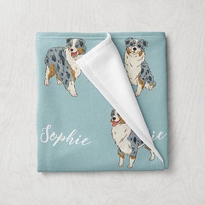 May include: A blue and white fleece blanket with a white border and a repeated pattern of Australian Shepherd dogs. The blanket is folded in half and the name "Sophie" is written in white cursive on the blanket.