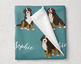 May include: A teal blue fleece blanket with a white fleece reverse side. The blanket features a repeating pattern of Bernese Mountain Dog illustrations and the name "Sophie" printed in white.