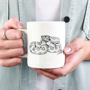 May include: White ceramic mug featuring a black and white illustration of a snake wearing glasses. The snake is coiled and has a detailed, patterned design. The mug is held by a person wearing a light green shirt.
