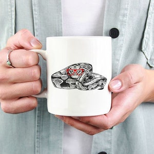 May include: White ceramic mug with a black and white illustration of a snake wrapped around itself. The snake's eyes are red.