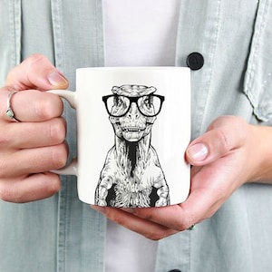 May include: White ceramic mug featuring a black and white illustration of a dinosaur wearing glasses. The dinosaur is looking directly at the viewer.