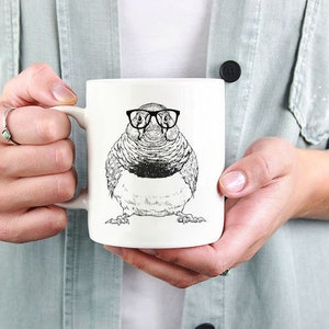 May include: White ceramic mug with a black and white illustration of a bird wearing glasses. The bird is round and has a short beak.