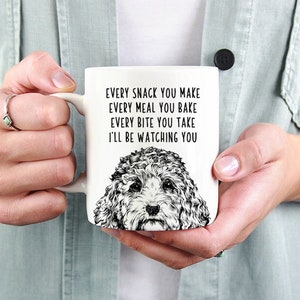 May include: White ceramic mug featuring a black and white illustration of a dog's face. The mug has the text "EVERY SNACK YOU MAKE, EVERY MEAL YOU BAKE, EVERY BITE YOU TAKE, I'LL BE WATCHING YOU" printed in black.