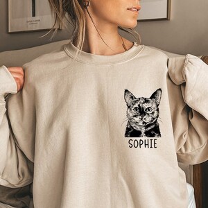 May include: A beige sweatshirt featuring a black and white illustration of a cat with the name "Sophie" printed on the front.