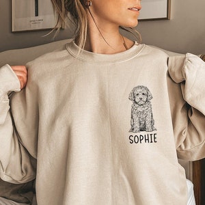 May include: Beige sweatshirt featuring a detailed black and white illustration of a dog, with the name "SOPHIE" printed below. The sweatshirt is a neutral color, and the dog illustration is centered on the chest.