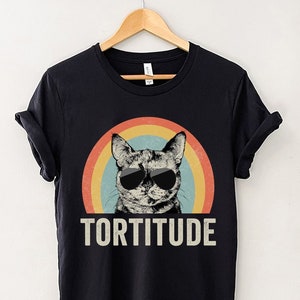May include: Black t-shirt with a retro rainbow design and a cat wearing sunglasses. The text "TORTITUDE" is printed below the cat.