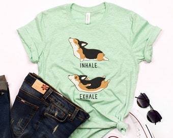 May include: A light green t-shirt with a graphic of a corgi dog in a yoga pose. The text "inhale" and "exhale" are printed below the dog. The shirt is paired with blue jeans, white sneakers, and black sunglasses.