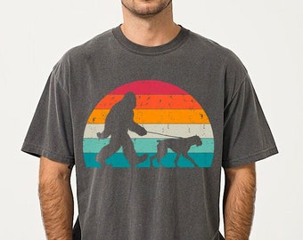 May include: Dark grey t-shirt with a Bigfoot silhouette walking a dog on a lead, set against a retro sunset design with orange, red, and teal stripes. The shirt has a relaxed fit.